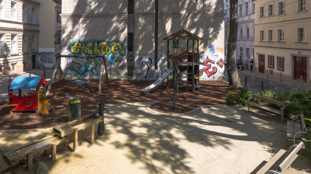 Spielplatz mit Graffiti in einer städtischen Umgebung, umgeben von Gebäuden, Bäumen, Bänken und einer Straße mit Menschen und Fahrzeugen.
