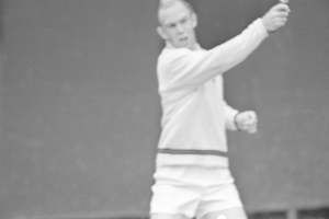 Ein Mann in weißer Tennisbekleidung steht auf einem Tennisplatz und hält einen Schläger in der Hand, mit entschlossener Miene, umgeben von grünem Gras unter einem strahlend blauen Himmel.