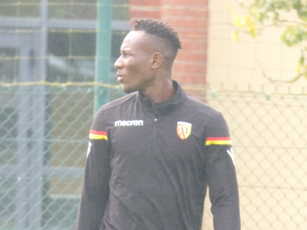 Ein Mann In Einer Schwarzen Jacke Steht Auf Einem Fußballfeld, Umgeben Von Einem Zaun Und Eines Baumes, Und Sieht konzentriert Aus During Einer Trainingseinheit.