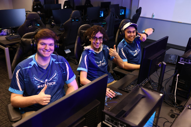 Drei junge Männer in Headsets lächeln, während sie an Computern mit Monitoren, Tastaturen und Kabeln sitzen, was darauf hindeutet, dass sie ein Spiel als Teil des Wake Tech Esports-Teams spielen.
