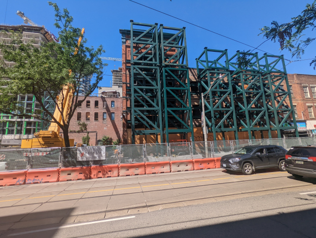 Eine Baustelle mit einer teilweise errichteten Metallbrücke im Vordergrund, parkende Autos am Straßenrand, ein Zaun, Bäume, Gebäude mit Fenstern, ein Kran und ein Himmel im Hintergrund.