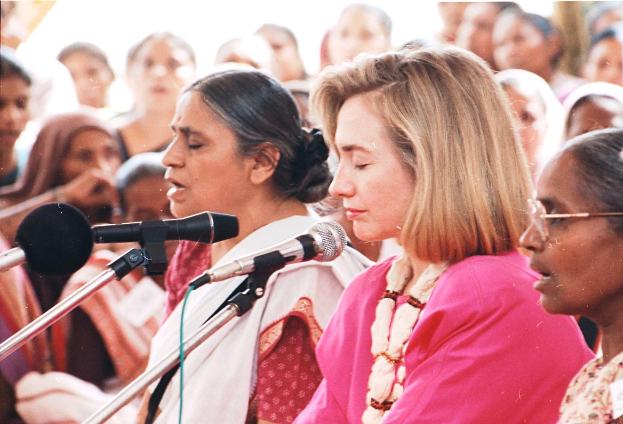 Eine Gruppe von Frauen steht vor einem Publikum, mit zwei Mikrofonen im Vordergrund und einer Frau, vermutlich Hillary Clinton, die die Menge anspricht.